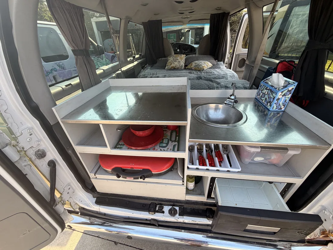 The rear-access kitchen: stainless prep counter, sink with faucet, pull-out propane stove, cutlery and storage, reached through the open back doors